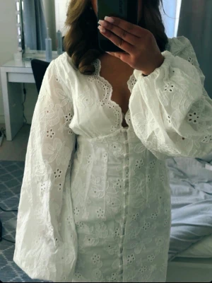Gina Tricot- Vit kort klänning med spets och knappar - Superfin vit kort klänning med broderade blommor, v-ringning och långa puffärmar. Klänningen har spetsdetaljer längs kanterna och knappar framtill. Perfekt för sommarens alla tillfällen!