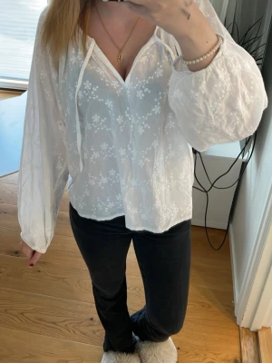 Vit blus med broderade blommor - Söt vit blus med broderade små blommor och ballongärmar. Blusen har v-ringning med knytband och är gjord i ett tunt, lätt material som ger en luftig känsla. 