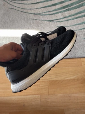 Svarta Adidas Ultraboost sneakers - Snygga svarta Adidas Ultraboost sneakers med stickad ovandel och vita Boost-sulor. Skorna har svarta snören och klassiska tre ränder på sidan. Perfekta för dig som gillar sportig och modern stil.