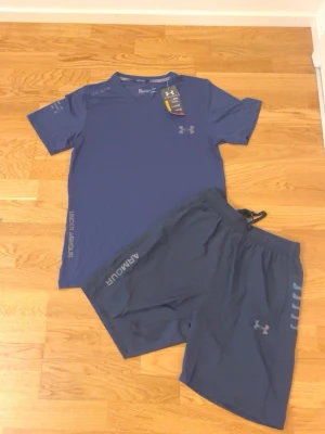 Blått träningsset från Under Armour - Säljer ett blått träningsset från Under Armour med t-shirt och shorts. Båda plaggen har diskreta loggor och är tillverkade i ett lätt och snabbtorkande syntetmaterial, perfekt för träning. T-shirten har korta ärmar och rund hals, shortsen har resår i midjan.