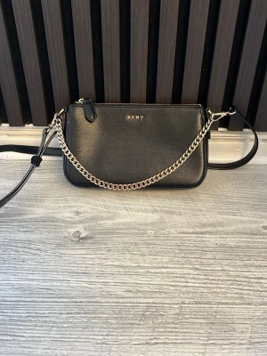 Svart axelväska från DKNY med kedja - Snygg svart axelväska från DKNY i slätt läder med silverfärgad kedja och justerbar axelrem. Väskan har minimalistisk design, dragkedja upptill och diskret DKNY-logga i guld framtill. Perfekt storlek för det viktigaste och stilren look. Axelrämmen går att ta bort ifall man vill använd den som en handväska 