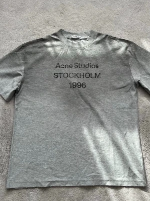 Grå Acne Studios t-shirt - Snygg grå t-shirt från Acne Studios med tryck på bröstet där det står 'Acne Studios STOCKHOLM 1996'. Klassisk passform och rund halsringning. Perfekt basplagg för en clean och stilren look.
