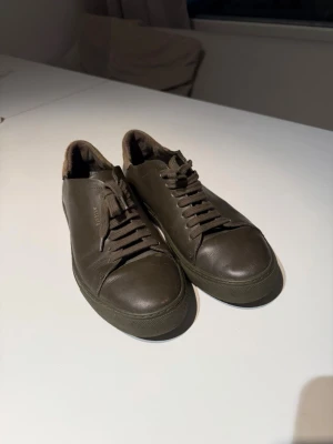 Olivgröna sneakers från Axel Arigato - Snygga olivgröna sneakers från Axel Arigato i slätt läder med matchande snörning och diskret logga på sidan. Låg modell med mjuk vadderad häl och klassisk gummisula. Perfekta för en clean och stilren look.