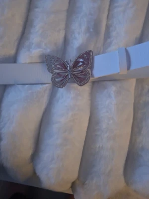 Vitt bälte med fjärilsspänne med 5 håll - Snyggt vitt bälte med ett detaljerat fjärilsspänne i silver och rosa. Bältet har en stilren design och passar perfekt för att ge din outfit en unik touch. Spännet har fina utskärningar och små stenar som ger extra bling. Det finns 5 spännes nivåer