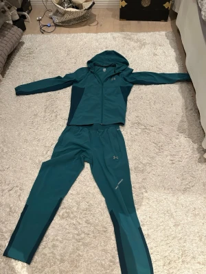 Turkos träningsset från Under Armour - Snyggt turkost träningsset från Under Armour med matchande hoodie och byxor. Setet har dragkedja på tröjan, huva och elastisk midja på byxorna. Materialet är lätt och stretchigt, perfekt för träning eller löpning. Mörkare detaljer längs sidorna för extra stil.