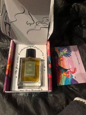 Sugar Leather Eau de Parfum - Unik parfym med namnet Sugar Leather i en elegant, rektangulär glasflaska med svart kork. Doften har en varm, bärnstensfärgad vätska och levereras i en konstnärligt designad förpackning. Perfekt för dig som gillar exklusiva och kreativa dofter.