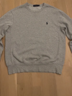 Grå sweatshirt från Polo Ralph Lauren - Klassisk grå sweatshirt från Polo Ralph Lauren med rund halsringning och diskret broderad logga på bröstet. Tröjan har ribbade muddar vid ärmslut och nederkant, perfekt för en clean och avslappnad stil.