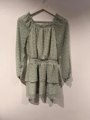 Grön blommig volangklänning - Söt ljusgrön klänning med små vita blommor, långa ärmar och volangdetaljer. Klänningen har resår i midjan och en luftig, flowy passform. Perfekt för vår och sommar, tillverkad i ett tunt och lätt tyg. Den har använts på en skolavslutning och är precis som ny. Den är från Kappahl i storlek 158. Material: Polyester.