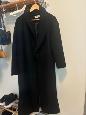 Svart ullkappa från Gina Tricot XS - Stilren svart kappa från Gina Tricot i ullblandning. Lång modell med klassisk krage och knäppning framtill. Perfekt för dig som vill ha en enkel och tidlös look. Passar till många olika outfits och håller dig varm under kyliga dagar. Bältet runt om är bortappat därav priset. Man kan köpa till ett bälte eller bara knäppa den.