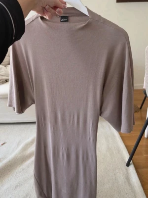Beige omlottklänning med knyt - Säljer en beige klänning i mjukt, tunt material med omlottdetalj och knyt i ryggen. Klänningen har korta ärmar och rak passform, perfekt för en avslappnad och stilren look.