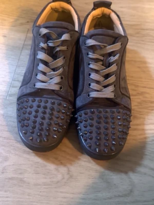 Louboutin skor - Unika svarta sneakers i mocka med grå skosnören och coola nitar över hela tån. Skorna har en klassisk låg profil och röd sula med signaturdetalj undertill. Perfekta för dig som vill sticka ut med din stil.