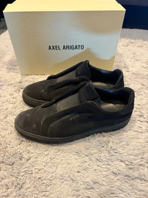 Svarta slip-on sneakers Axel Arigato - Stilrena svarta slip-on sneakers från Axel Arigato i mocka och textil. Diskret logga på sidan och tungan. Skorna har en låg profil och minimalistisk design utan snörning, perfekt för en clean och modern look.