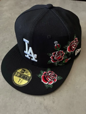 Helt ny new era keps med unik design - Snygg svart keps från New Era med LA-logga framtill och broderade röda rosor på sidan och skärmen. Klassisk 59FIFTY-modell med rak skärm och broderade detaljer i vitt, rött och grönt. Perfekt för dig som vill sticka ut med en unik accessoar.