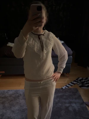 Vit blus med volangkrage och rosett - Söt vit blus med transparenta långärmar, stor volangkrage och dekorativ svart rosett i halsen. Blusen har en romantisk och vintage-inspirerad look med fina spetsdetaljer på kragen. Perfekt för dig som gillar en feminin och trendig stil.