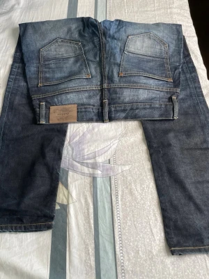 Mörkblå raka jeans med slitningar - Säljer ett par mörkblå jeans med raka ben och snygga slitningar framtill. Jeansen har klassiska fem fickor och kontrastsömmar. Materialet är denim i bomull och passformen är avslappnad men ändå stilren.