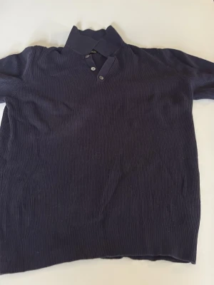 Mörkblå ribbad polotopp Massimo Dutti - Snygg mörkblå ribbad polotopp från Massimo Dutti i bomull och linne. Toppen har korta ärmar, krage och knappar framtill för en klassisk look. Perfekt till jeans eller linnebyxor för en stilren outfit.