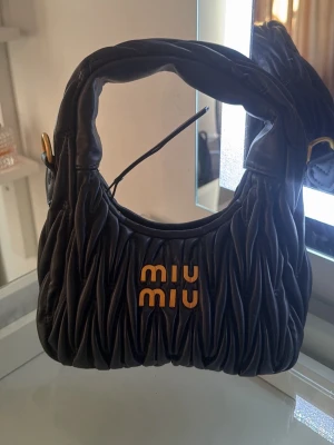 Svart handväska  - Trendig svart handväska  i mjukt, rynkat läder med guldfärgade detaljer och logotyp framtill. Väskan har en rundad form och kort axelrem, perfekt för att lyfta vilken outfit som helst. 