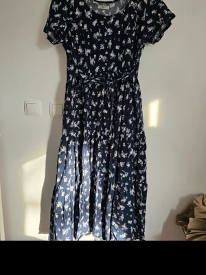 Mörkblå blommig långklänning - Söt långklänning i mörkblått med vitt och rosa blommönster. Klänningen har korta ärmar, rund halsringning och ett knytband i midjan för en feminin siluett. Perfekt för varma dagar och har en luftig, flowy passform.