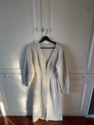 Ljusbeige lång stickad klänning - Mysig lång stickad klänning i ljusbeige ton med v-ringning och knappar framtill. Klänningen har långa ärmar och ribbad struktur, perfekt för kyligare dagar. Enkel och stilren design som passar till många olika looks.