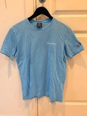 Ljusblå Champion t-shirt - Snygg ljusblå t-shirt från Champion med klassisk logga broderad på bröstet och ärmen. Tillverkad i mjuk bomull och har en normal passform med korta ärmar och rund hals. Perfekt för en avslappnad och sportig stil.