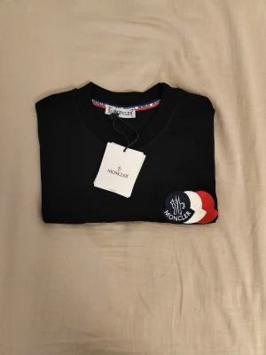 Svart Moncler t-shirt med logga - Snygg svart t-shirt från Moncler med klassisk rund hals och korta ärmar. På bröstet sitter Monclers ikoniska logga i rött, vitt och blått. Tillverkad i mjuk bomull för en skön känsla. Perfekt till jeans eller shorts.