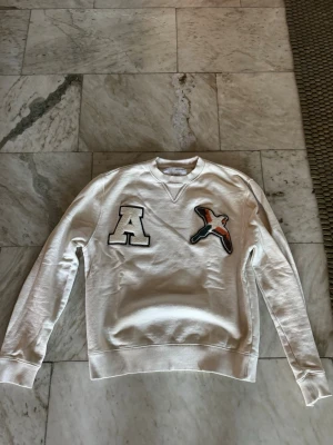 Beige sweatshirt Axel Arigato - Vit sweatshirt från Axel Arigato med broderad bokstav 'A' och en färgglad fågel på bröstet. Tröjan har rund halsringning, ribbade muddar och är tillverkad i mjukt material. Perfekt för en avslappnad och trendig stil. Storlek XS. Kontakta mig vid frågor!😃
