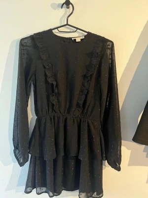 Svart volangklänning med prickar - Söt svart kortklänning med diskreta prickar och volangdetaljer framtill. Klänningen har långärmade transparenta ärmar och rund halsringning. Tillverkad i ett lätt och luftigt material som ger en flowy känsla.