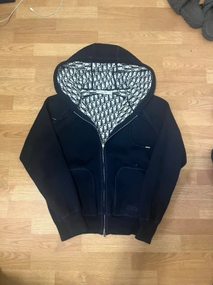 Svart  hoodie med monogramfoder - Säljer en svart hoodie från Di@r med dragkedja och fickor framtill. Insidan av huvan har ett snyggt svartvitt Di@r-monogrammönster och loggan är diskret broderad på ena fickan. Perfekt för dig som gillar exklusiva streetwear-plagg.
