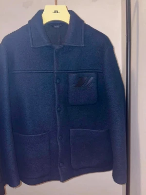 Mörkblå ull overshirt från J.Lindeberg - Stilren mörkblå ull overshirt från J.Lindeberg i ullmix. Klassisk krage, tre framfickor och diskret logga på bröstfickan. Perfekt lager-på-lager-plagg med knappstängning och avslappnad passform.