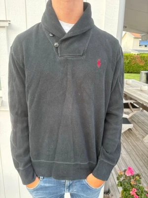 Mainblå tröja med sjalkrage från Polo Ralph Lauren - Snygg marinblå kofta från Polo Ralph Lauren med sjalkrage och två knappar vid halsen. Tröjan har lång ärm, ribbade muddar och den klassiska röda loggan broderad på bröstet. Perfekt för en chill och stilren look.