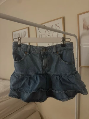 Blå volangkjol i denim - Säljer en blå jeanskjol med volang och klassisk femficksdesign. Perfekt för dig som gillar en avslappnad och trendig stil. 