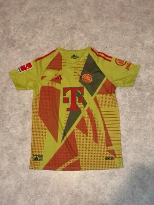 Bayern München målvaktströja gul/röd - Snygg Bayern München målvaktströja i gult med röda och gröna grafiska detaljer. Adidas-logga, klubbmärke och sponsortryck på framsidan. Nummer 1 och Neuer på ryggen. Korta ärmar och ventilerande material, perfekt för fotbollsträning.