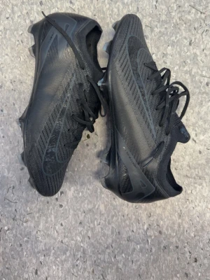 Svarta fotbollsskor med dobbar - Säljer ett par helsvarta fotbollsskor med snörning och låg profil. Skorna har en modern, strömlinjeformad design med mönstrad ovandel och fasta dobbar för bra grepp på planen. Tillverkade i syntetmaterial och mesh för lätt känsla och bra ventilation.