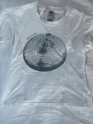 Vit t-shirt med discokula-tryck - Säljer en vit t-shirt med ett stort tryck av en discokula i svart på framsidan. T-shirten har rund hals och korta ärmar. Perfekt för dig som gillar coola och unika prints. Materialet känns mjukt och skönt, troligtvis bomull.