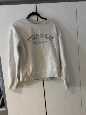 Grå sweatshirt med Venice-tryck XS - Ljusgrå sweatshirt med texten 'VENICE Caffè Panorama' broderat framtill i mörkblått och vitt. Tröjan har rund hals, ribbade muddar vid ärmslut och nederkant samt en avslappnad passform. Perfekt för en chill och casual stil.