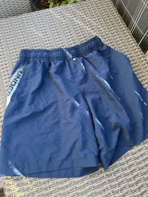Blå träningsshorts från Under Armour - Snygga marinblå träningsshorts från Under Armour med elastisk midja och tryckt logga längs sidan. Perfekta för gymmet eller löprundan. Lätta och luftiga med avslappnad passform.
