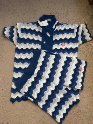 Blåvitt vågmönstrat set med shorts Från CasaBlanca - Säljer ett snyggt tvådelat set med kortärmad topp och shorts i blått, vitt och ljusblått vågmönster. Setet har dekorativa knappar framtill, krage och matchande vågiga kanter. Materialet är stickat och mjukt, perfekt för en trendig look.