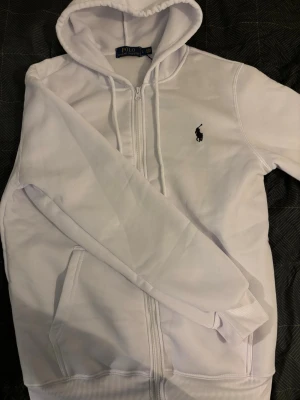 Vit hoodie från Polo Ralph Lauren - Snygg vit hoodie med dragkedja från Polo Ralph Lauren. Klassisk design med huva och dragsnören, två fickor framtill och den ikoniska broderade loggan på bröstet. Tillverkad i mjukt bomullsmaterial för maximal komfort.