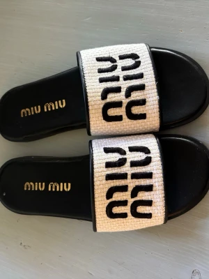 Svarta sandaler från Miu Miu - Snygga svarta sandaler från Miu Miu med bred vit rem i textil och svart logotyp. De är inte äkta!! Äkta skorna kostar ca 8000kr. Bara använda ca 3 gånger