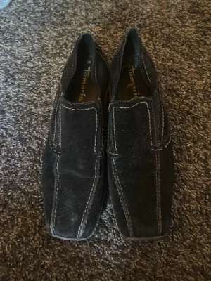 Svarta mockaloafers från Tamaris - Säljer ett par svarta loafers från Tamaris i mjuk mocka med kontrasterande vita sömmar. Skorna har fyrkantig tå och en låg sula i gummi som ger en clean look. Perfekta för dig som gillar klassisk men ändå trendig stil.
