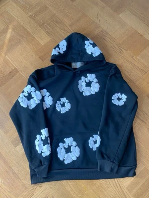 Svart hoodie med vita blommor - Säljer en svart hoodie med huva och stora vita blommönster tryckta över hela tröjan. Hoodien har en klassisk känguruficka framtill och ribbade muddar vid ärmar och nederkant. Perfekt för dig som gillar streetwear och vill sticka ut.