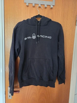 Svart Sail Racing hoodie med tryck - Snygg svart hoodie från Sail Racing med vitt tryck på bröstet och logga på huvan. Klassisk känguruficka framtill och justerbar dragsko i huvan. Tillverkad i mjukt bomullsmaterial, perfekt för en avslappnad stil.