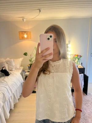 Vit ärmlös broderad topp - Söt vit ärmlös topp med broderade blommor och hålmönster. Rund halsringning och knapp i nacken för stängning. Köpt begagnad så vet ej vilket varumärke eller storlek, men skulle säga XS/S