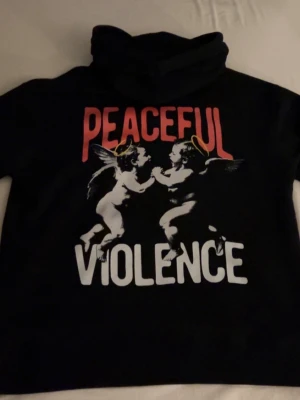 Svart hoodie från MANTO - Säljer en svart hoodie från MANTO. På baksidan finns ett stort tryck med två änglar och texten 'PEACEFUL VIOLENCE' i rött och vitt. Framsidan har en enkel vit MANTO-logga. Perfekt för dig som gillar streetwear och unika prints. Tröjan är lite stor i storlek. Aldrig använd!