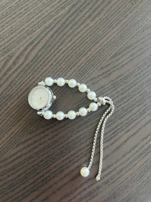 Pärlarmbandsklocka i silverfärg och vitt - Stilren klocka med armband av vita pärlor och silverfärgade detaljer. Urtavlan är rund och vit med minimalistiska markeringar och silverram. Justerbart armband med pärldetalj i änden för en elegant look. Saknar en pärla, men egentligen inget man tänker på enligt mig. Justerbar