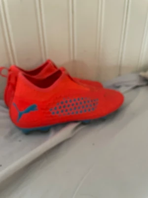 Röda Puma fotbollsskor med blå detaljer - Säljer ett par orange/röda fotbollsskor från Puma med blå sula och blått mönster på sidorna. Skorna har låg profil, strumpliknande passform och är tillverkade i syntetmaterial med meshpaneler för extra ventilation. Perfekta för dig som vill sticka ut på planen.
