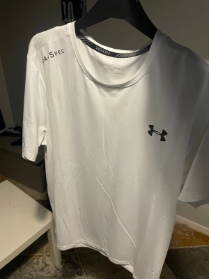 Vit tränings-tshirt från Under Armour - Säljer en vit tränings-tshirt från Under Armour med rund hals och korta ärmar. Tillverkad i ett lätt och snabbtorkande funktionsmaterial, perfekt för löpning eller gym. Diskret logga på bröstet och textdetalj på axeln.