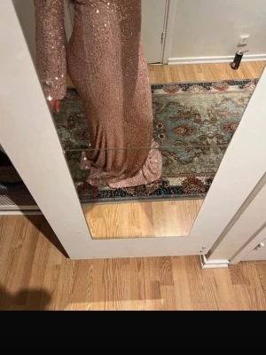 Lång glittrig rosa klänning - Säljer en elegant långklänning i ljusrosa med glittriga paljetter över hela plagget. Klänningen har lång ärm och figurnära passform som ger en snygg siluett. Perfekt för dig som vill sticka ut på bal eller fest.