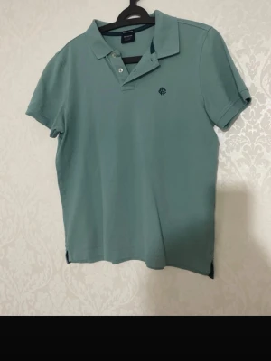 Grön pikétröja med krage - Snygg grön pikétröja med klassisk krage och korta ärmar. Tröjan har knappar vid halsen och en liten broderad logga på bröstet. Perfekt till jeans eller shorts för en avslappnad stil.
