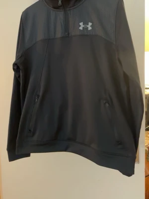Svart half-zip hoodie från Under Armour - Snygg svart hoodie från Under Armour med half-zip framtill och två dragkedjeförsedda fickor. Loggan sitter på bröstet och materialet är mjukt och sportigt, perfekt för chill eller träning. Regular fit och stilren design.